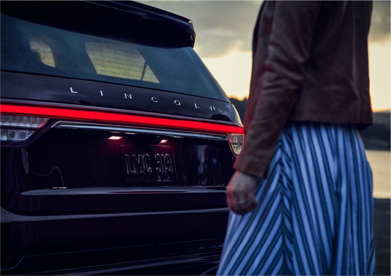 A person is shown near the rear of a 2023 Lincoln Aviator® SUV as the Lincoln Embrace illuminates the rear lights | Joe Cooper Lincoln of Edmond in Oklahoma City OK
