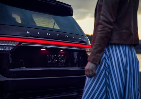 A person is shown near the rear of a 2024 Lincoln Aviator® SUV as the Lincoln Embrace illuminates the rear lights | Joe Cooper Lincoln of Edmond in Oklahoma City OK
